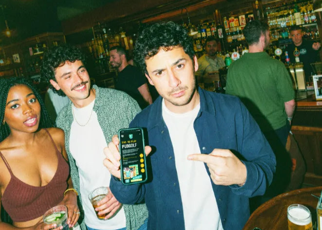 Friends in a pub holding up a phone with the Pub Golf Club app, pints on the table