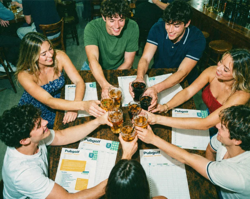 Group of eight clinking pints around a pub table with their pub golf scorecards laid out
