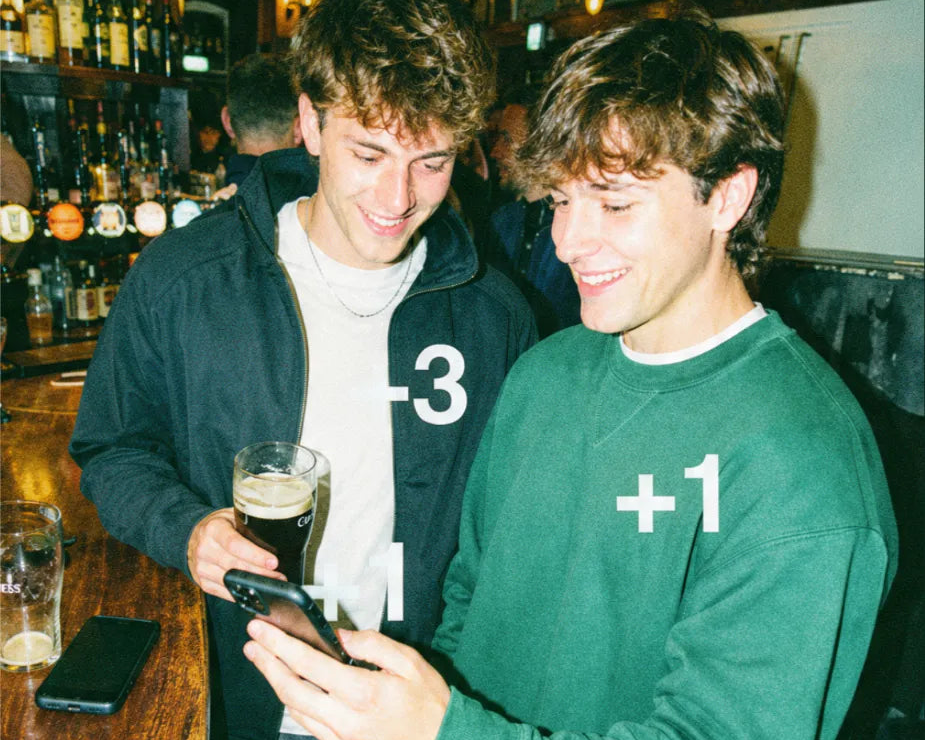 Two players checking their live pub golf scores on a phone at the bar, marked minus three and plus one