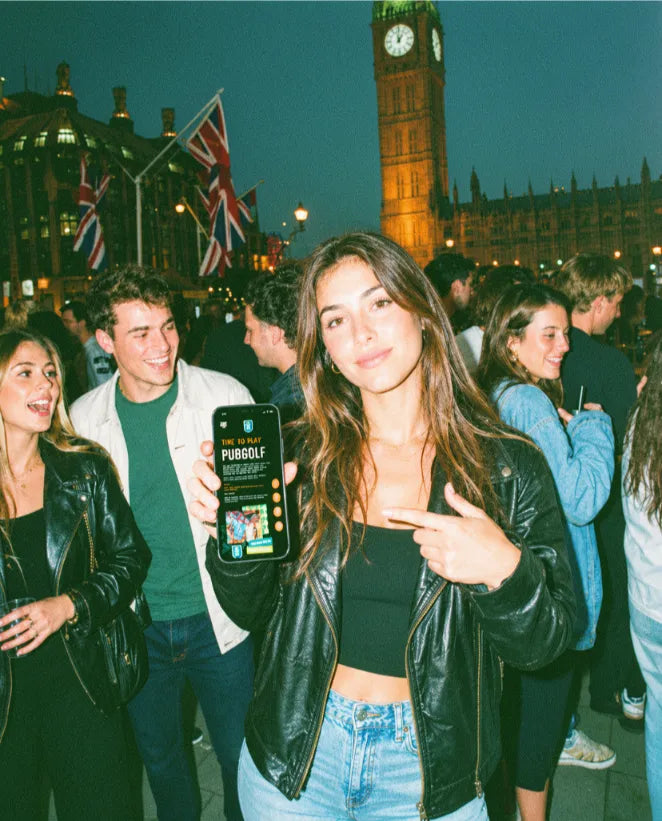 Young woman holding up a phone with the Pub Golf Club app in front of Big Ben at night in London