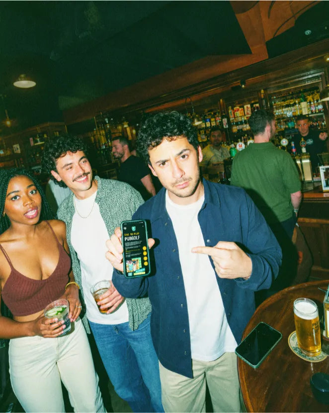 Friends in a pub holding up a phone with the Pub Golf Club app, pints on the table