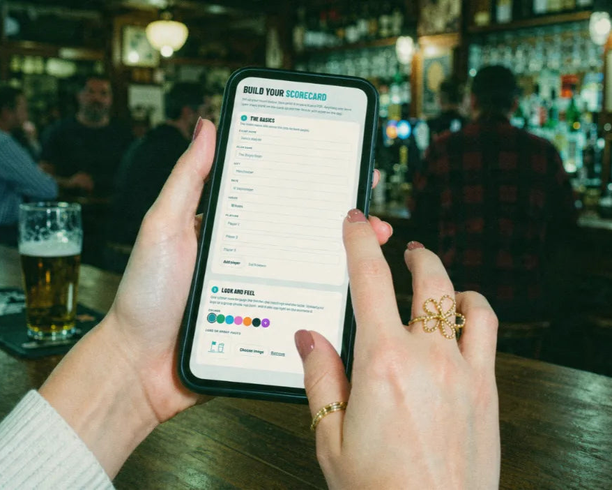 Hand holding a phone with the Build Your Scorecard form open, setting event name, city and players