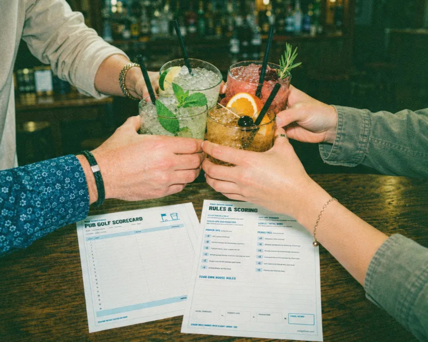 Three friends clinking colourful cocktails over their printable pub golf scorecard and rules sheet