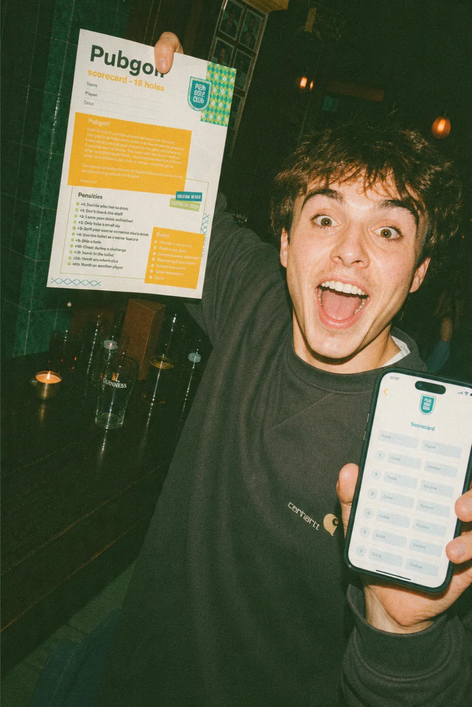 Player holding up an 18 hole pub golf scorecard beside the Pub Golf Club app open on a phone